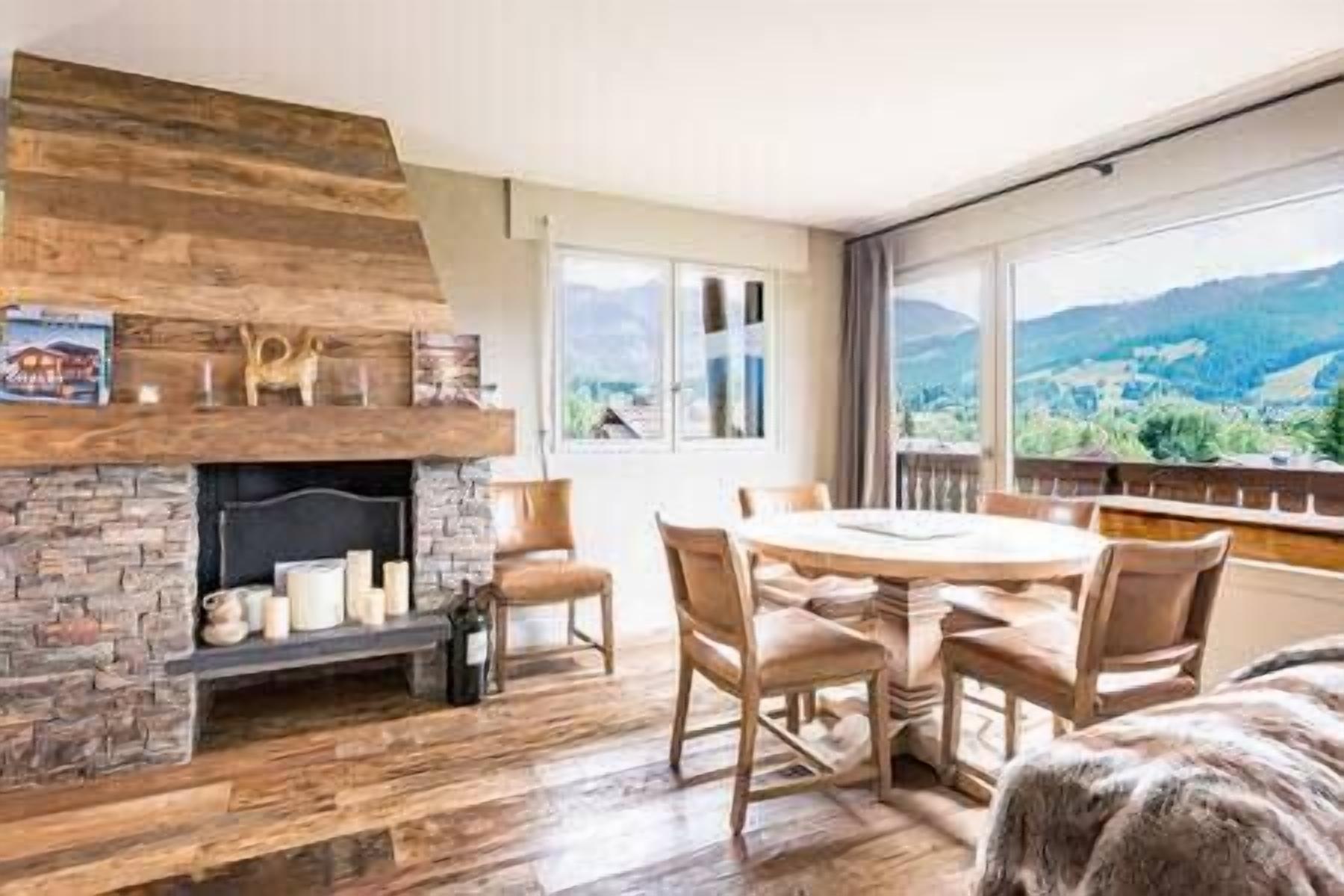 A room with a stone fireplace, wooden flooring, a round table with chairs, and a view of mountains through large windows.