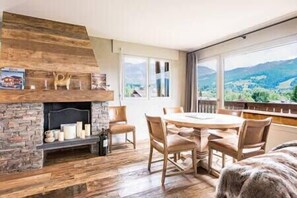 Ein Zimmer mit Kamin aus Stein, Holzboden, einem runden Tisch mit Stühlen und Bergblick durch große Fenster.