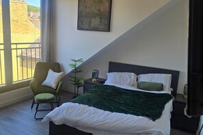 A bedroom with a bed, a chair, a nightstand, a lamp, and a window with a view of buildings. - Studio - Sleeps 2- Parking (Sheffield)