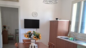 A dining area with a wooden table and chairs, a vase with sunflowers, a wall-mounted TV, and a kitchen with a sink and cabinets. - The Residences 6 (Marina di Modica)