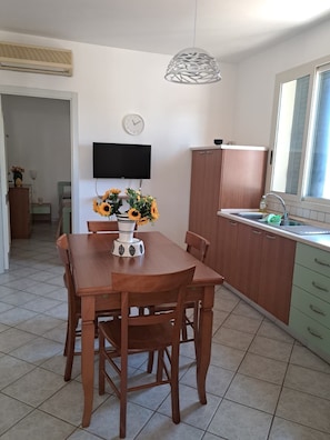 A dining area with a wooden table and chairs, a vase with sunflowers, a wall-mounted TV, and a kitchen with a sink and cabinets. - The Residences 6 (Marina di Modica)