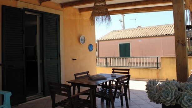 A covered outdoor patio with a wooden table and chairs, a hanging light fixture, and a view of a building with a tiled roof. - The Residences 6 (Marina di Modica)