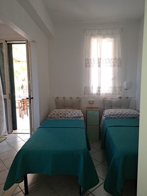 A room with two beds, a small bedside table, a window with curtains, and a door leading to an outdoor area. - The Residences 6 (Marina di Modica)