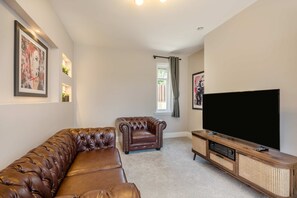A living room with a brown leather sofa, a brown armchair, a television on a wooden stand, and a window with curtains. - Detached Coastal Home - Walk to Beach - Sleeps 4 (Overstrand)