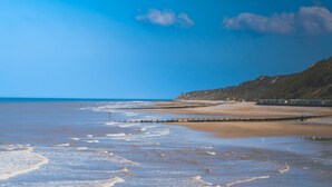 Ein Küstenstrand mit einer hölzernen Mole, die ins Meer führt, einem sandigen Ufer und einer Klippe im Hintergrund.
