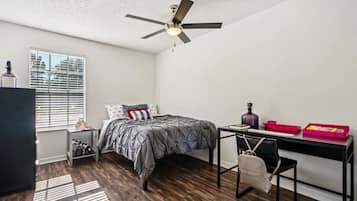 A bedroom with a bed, a desk, a chair, a ceiling fan, a window with blinds, and a radiator.