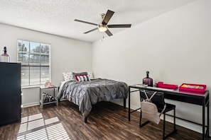 A bedroom with a bed, a desk, a chair, a ceiling fan, a window with blinds, and a radiator.