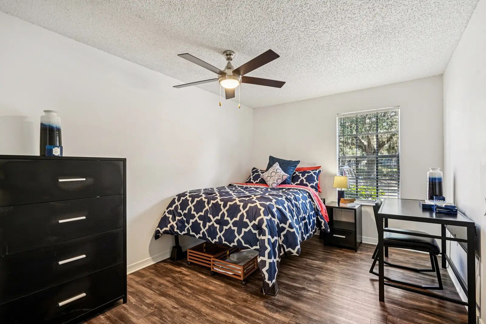 A bedroom with a bed, dresser, desk, and window.