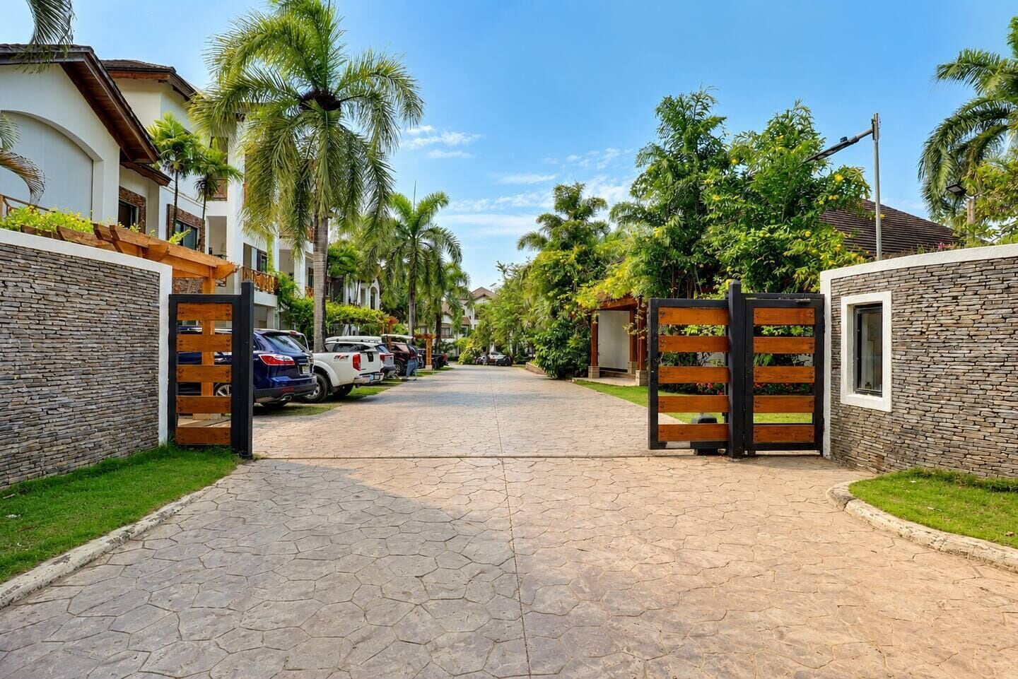 A gated driveway leading to a residential area with parked cars and palm trees.