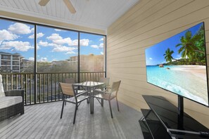 Ein Balkon mit Tisch und Stühlen, einer Couch und einem großen Fernseher, der eine Strandansicht zeigt.