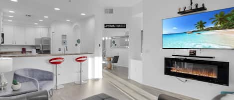A modern living room with a flat-screen TV, a fireplace, and a kitchen in the background.