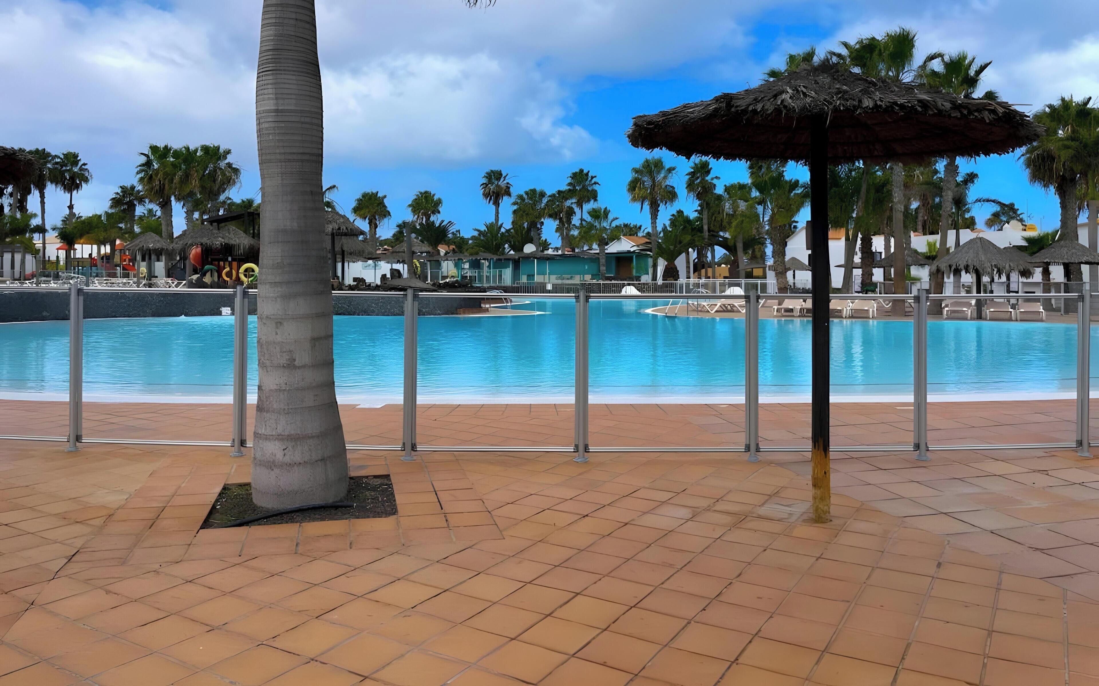 Una piscina con borde de baldosas, palmeras y una sombrilla de paja.