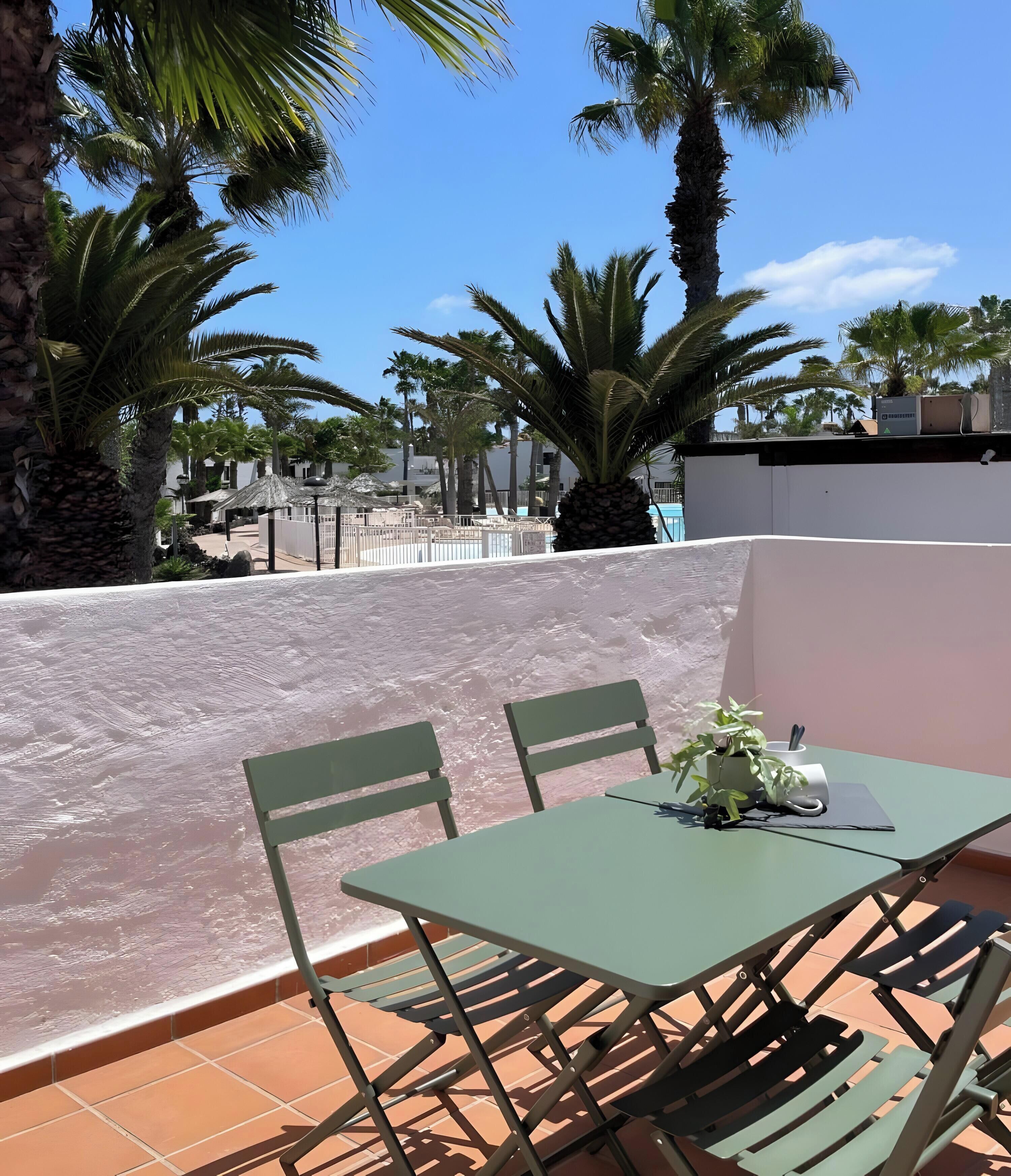 Eine Dachterrasse mit Tisch und Stühlen, umgeben von Palmen und blauem Himmel.