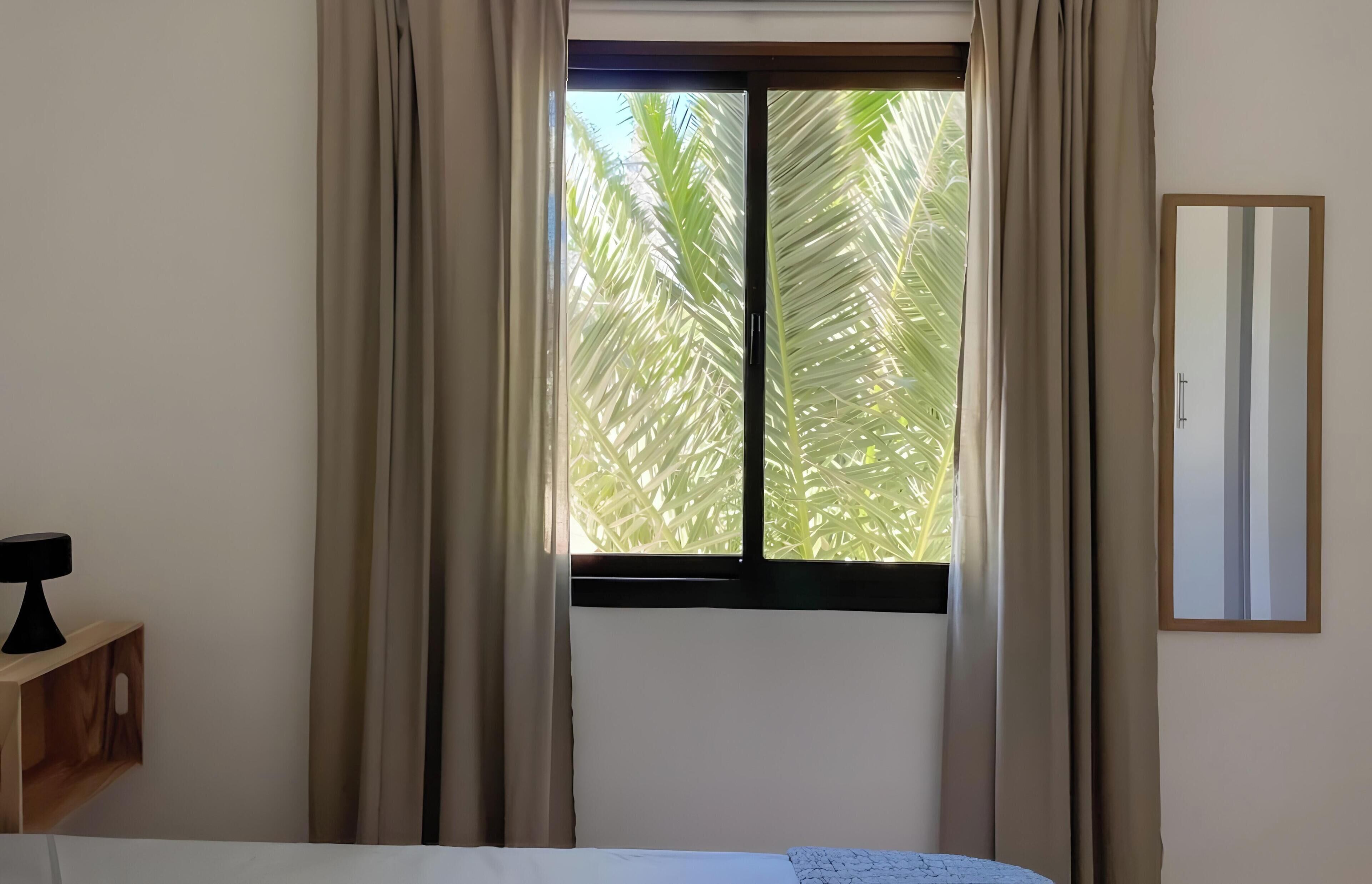 Una habitación con cama, mesita de noche, ventana con cortinas y espejo adosado a la pared.