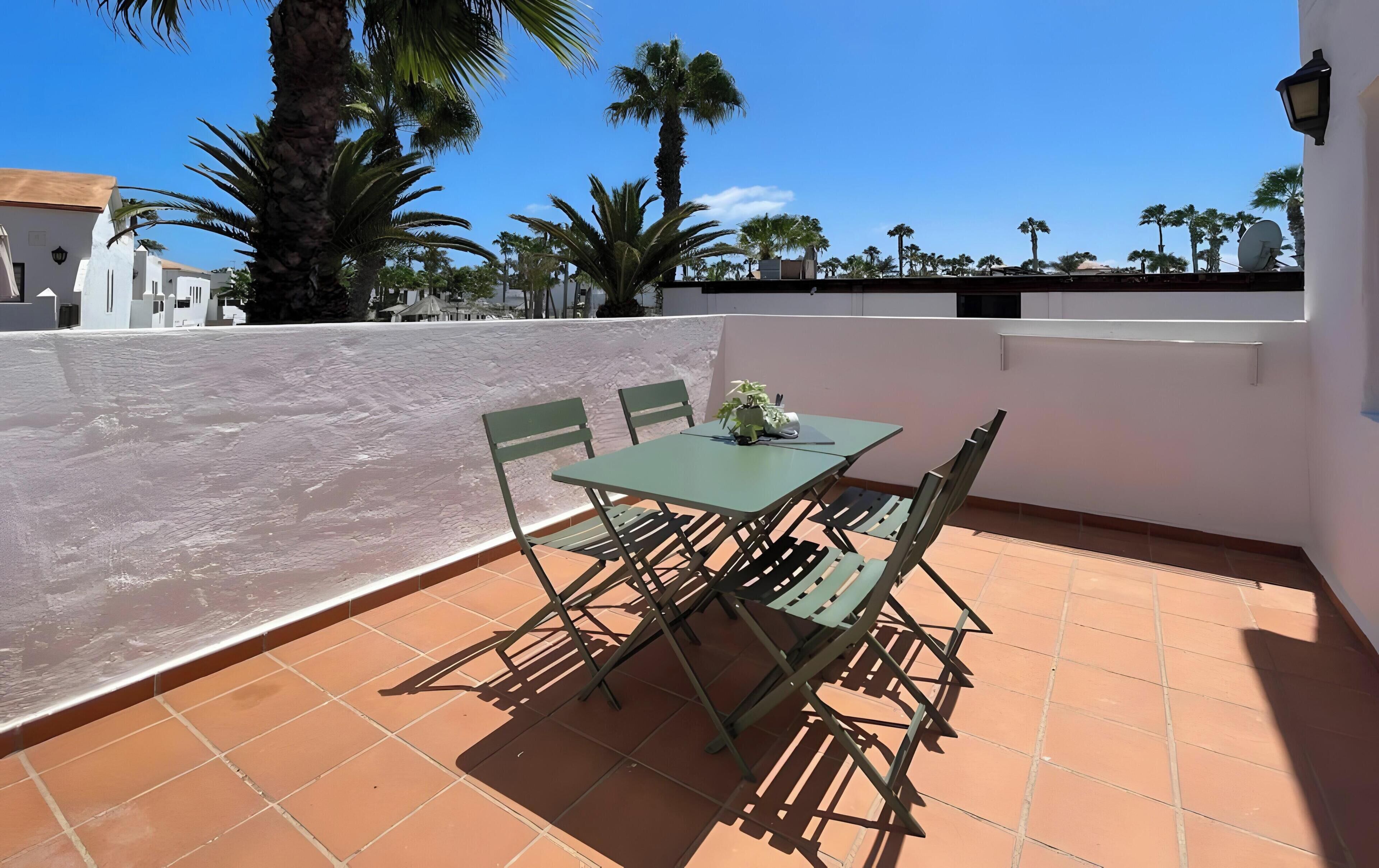 Una terraza con una mesa y sillas, una planta en maceta y vistas a palmeras y edificios.