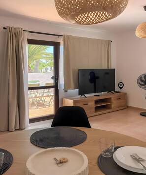 A modern living room with a television, wooden entertainment unit, dining table with placemats, and a sliding glass door leading to an outdoor area. - Oasis Dunas Canary House Deluxe by B-Rent – Luxury & Comfort in Corralejo (Corralejo)