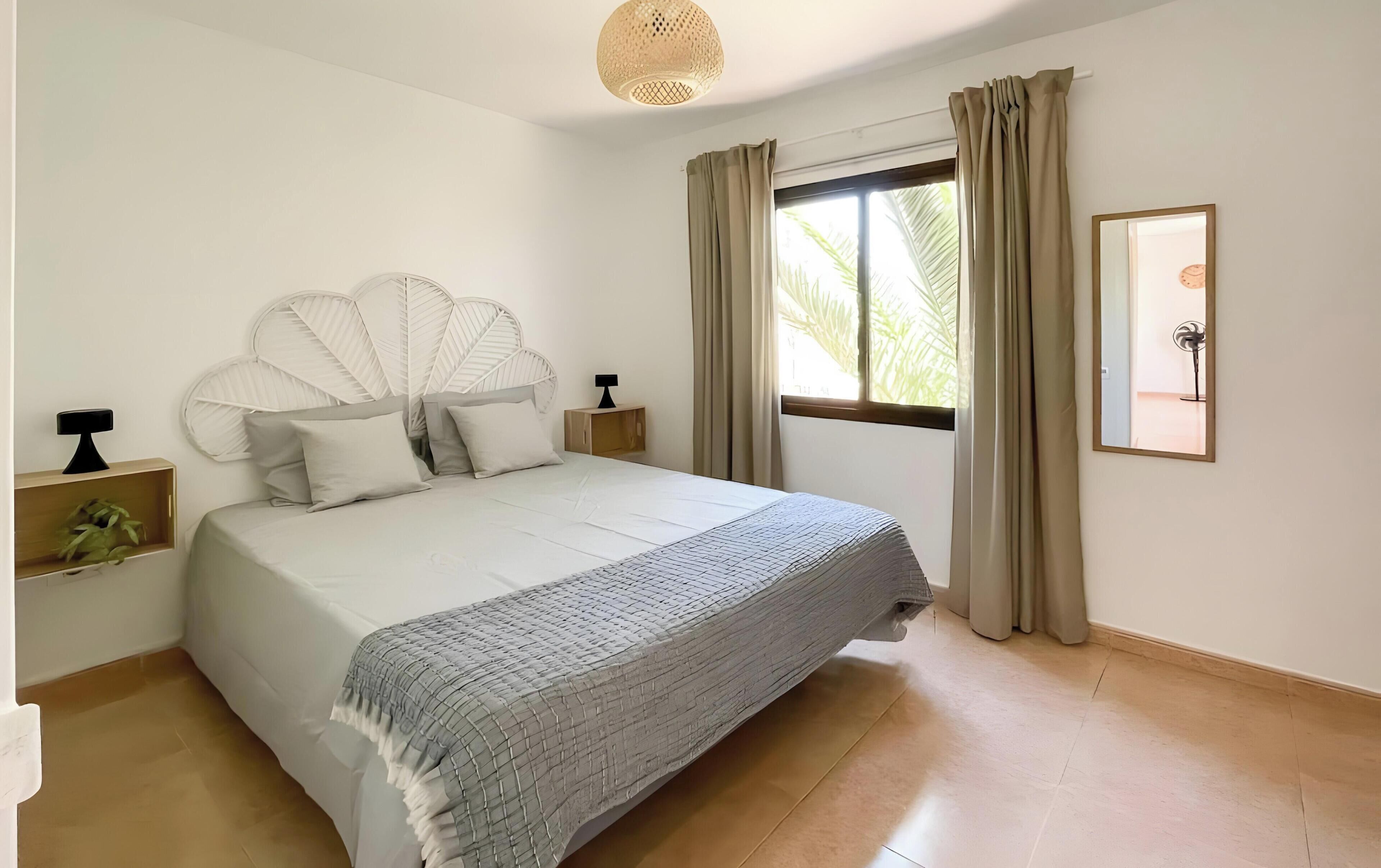 Un dormitorio ordenado con cama, mesitas de noche, una ventana con cortinas y una lámpara fijada a la pared.