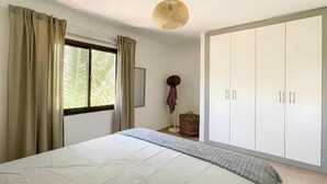 Ein Schlafzimmer mit Bett, Kleiderschrank, Pendelleuchte und einem Fenster mit Vorhängen.