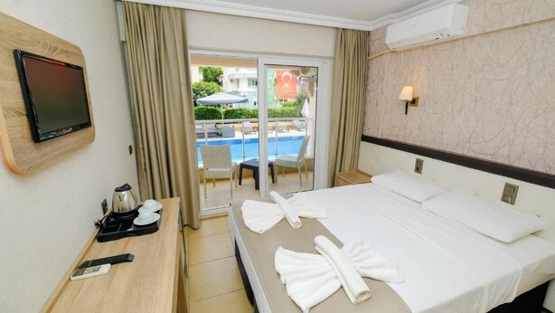 A hotel room with a bed, a desk with a kettle, a remote control, and a TV mounted on the wall. - MONİ HOTEL (Marmaris)
