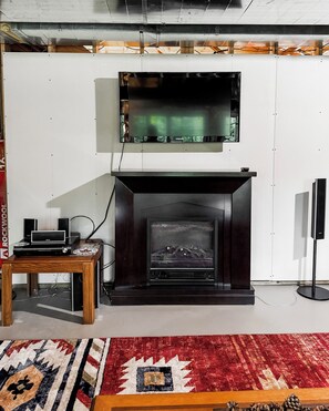 Ein Wohnzimmer mit einem Flachbildfernseher über einem Kamin, einem Couchtisch aus Holz und elektronischen Geräten sowie einem gemusterten Teppich auf dem Boden.