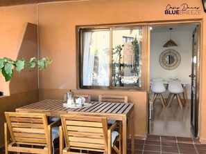 A wooden dining table with chairs, a window with curtains, and a view of the outside. - Oasis Dunas Blue Breeze Corralejo by B-Rent – Garden View Apartment (Corralejo)