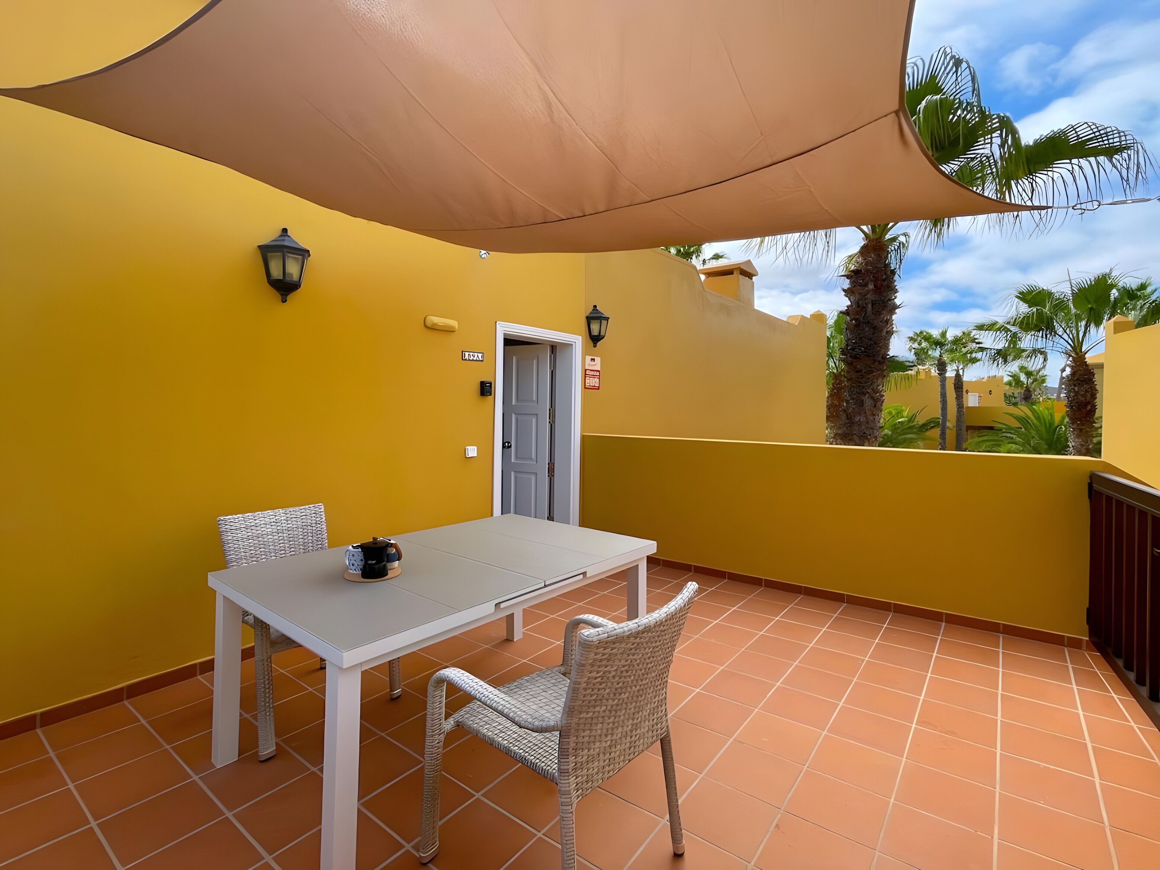 Un balcón con una mesa blanca y sillas de mimbre, un dosel color beige, y vistas a palmeras y un cielo despejado.