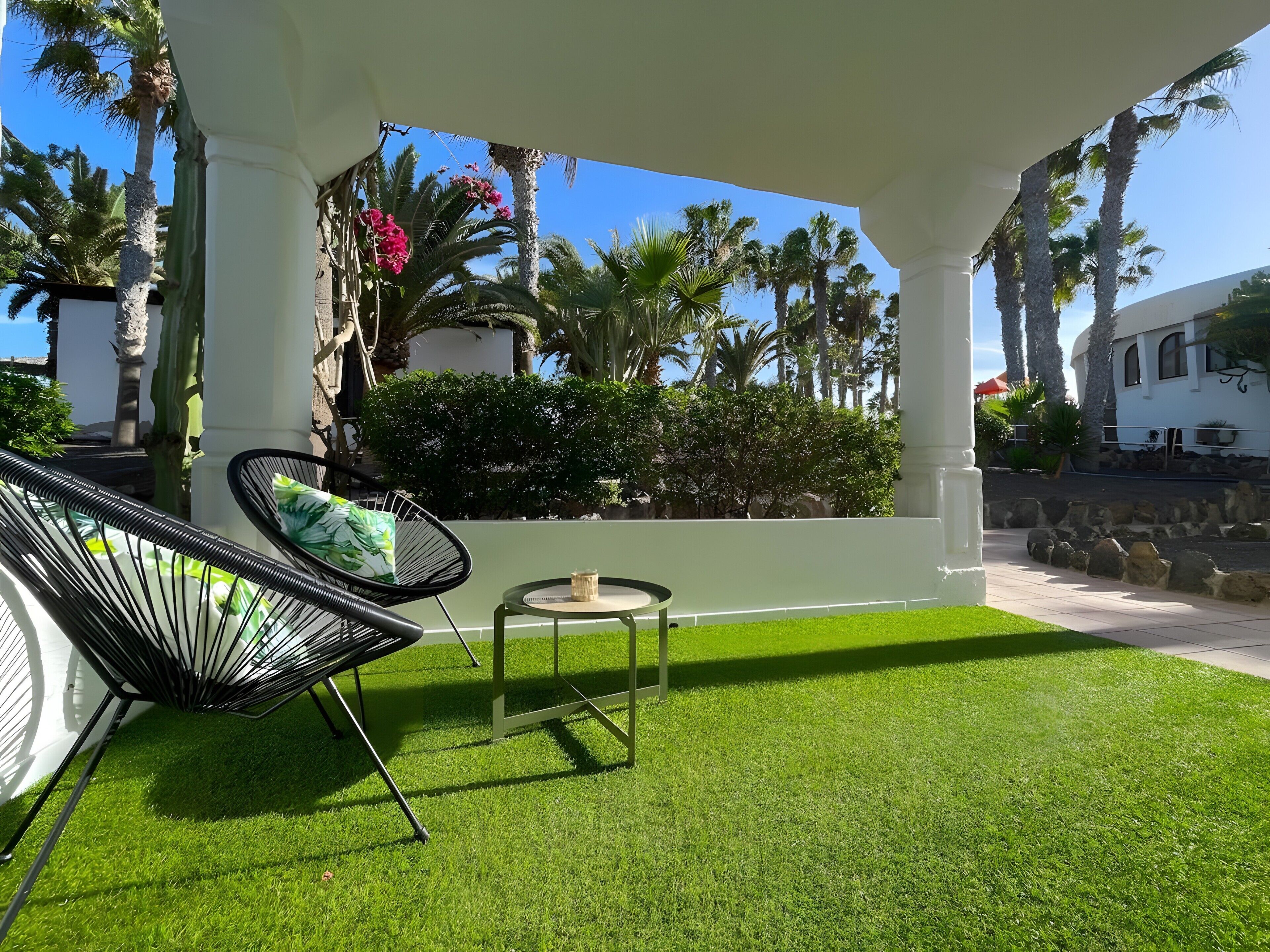 Un espace extérieur aménagé, abrité et doté de chaises et d’une table, entouré de palmiers et d’une pelouse bien entretenue.