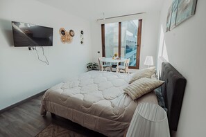 A bedroom with a bed, a TV mounted on the wall, a dining table with chairs, and a window with a view. - Executive Office · Gym · Coworking Space · Cumming Metro Station (Santiago)