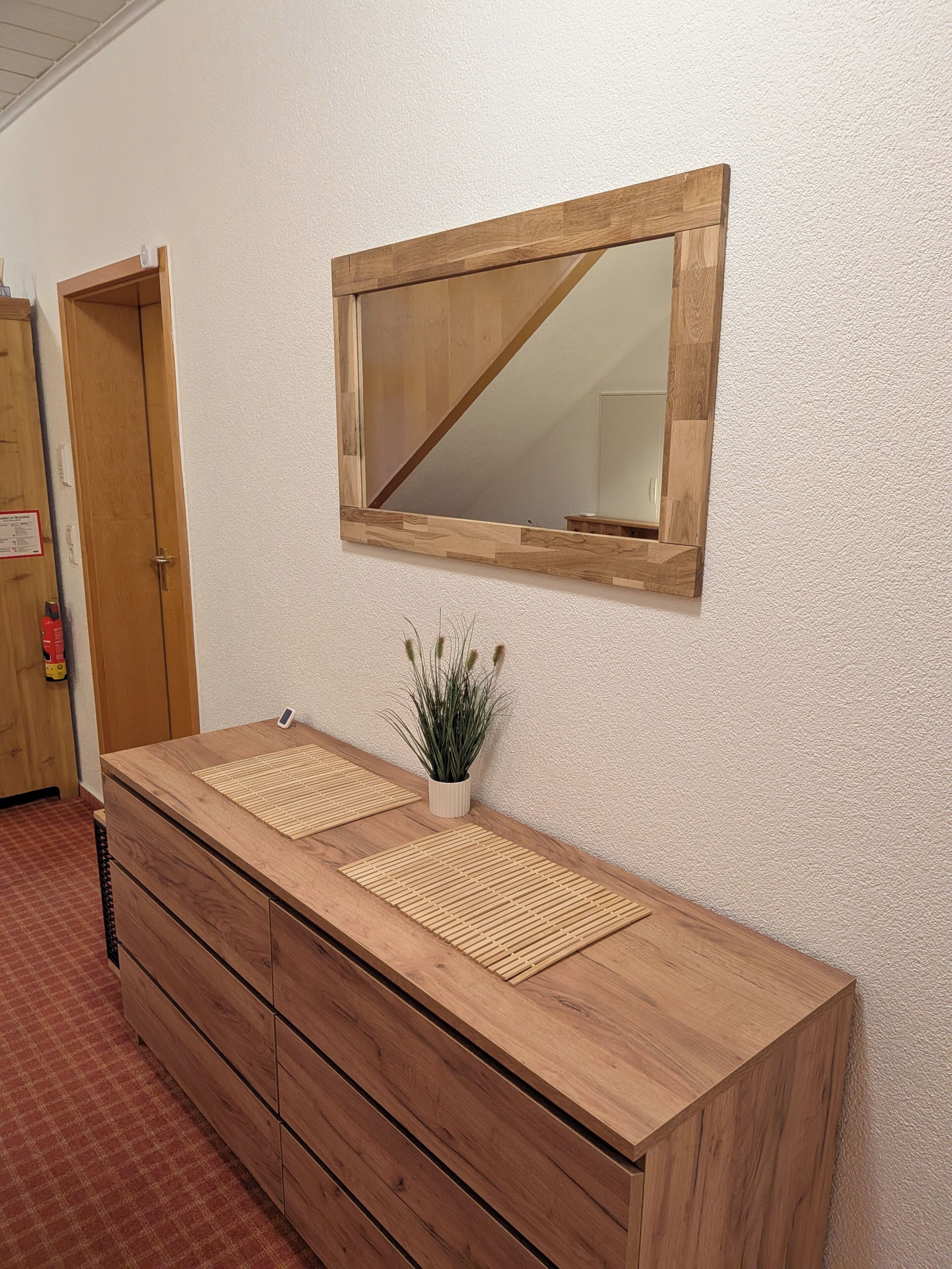 Ein hölzerner Waschtisch mit Spiegel darüber, eine kleine Topfpflanze und eine karierte Matte auf der Arbeitsfläche.