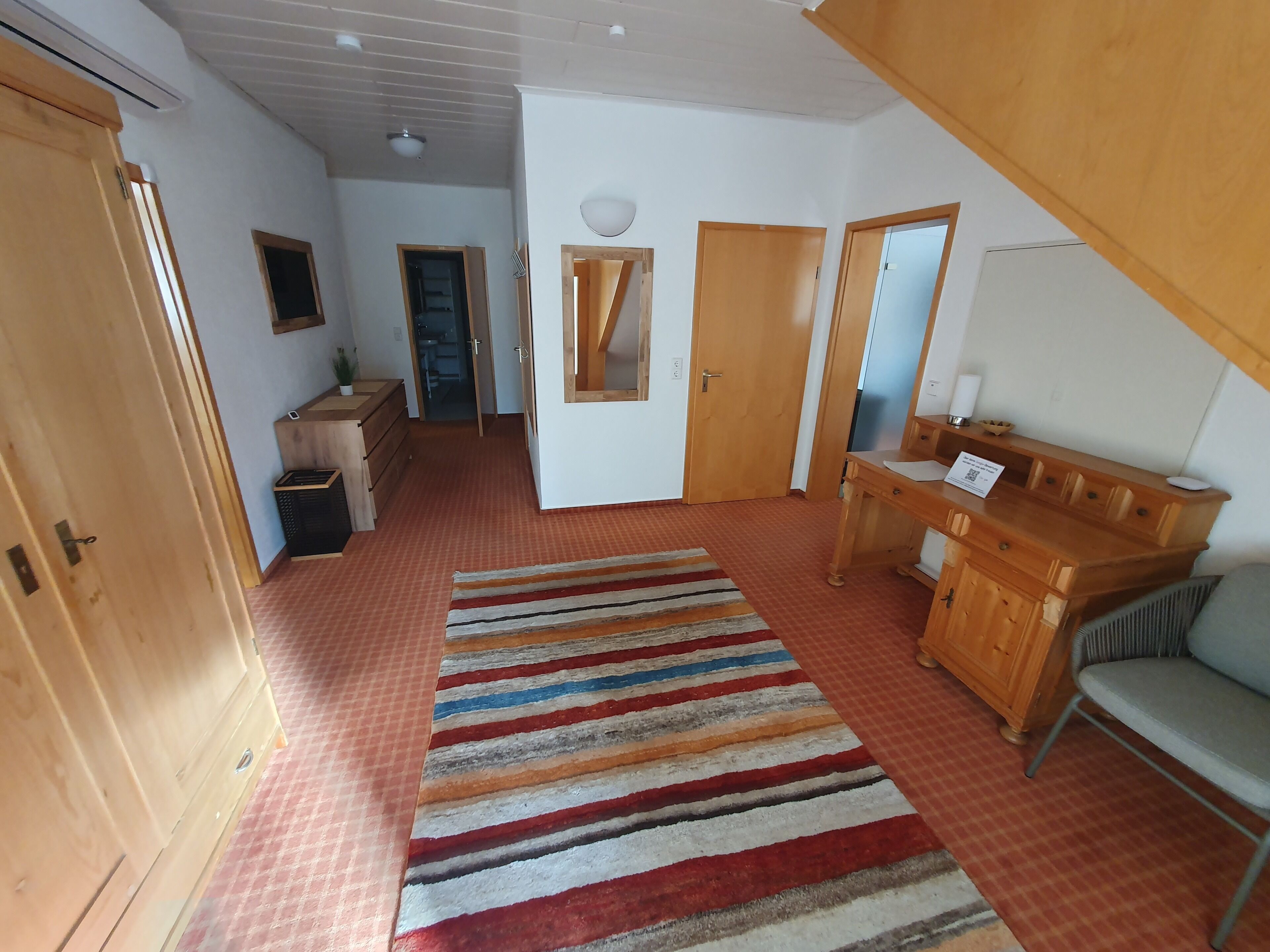 Ein Zimmer mit einem Holzschrank, einem Schreibtisch mit Spiegel, einem Streifenmuster-Teppich und einer Tür zu einem weiteren Raum.
