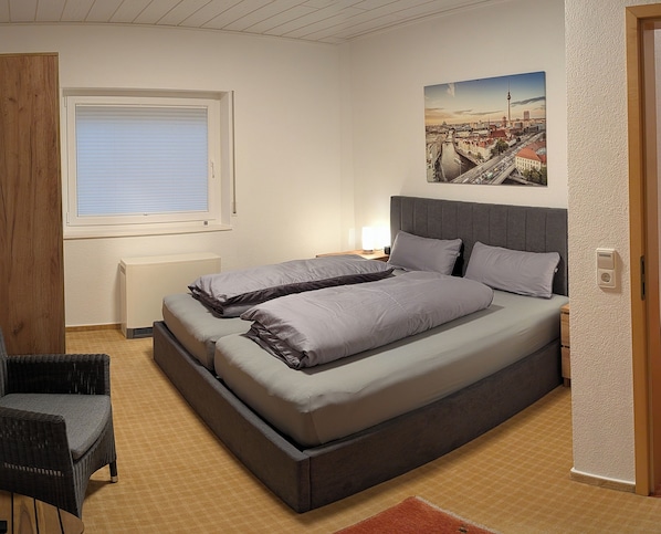 A bedroom with a bed, a chair, a window, and a cityscape painting.