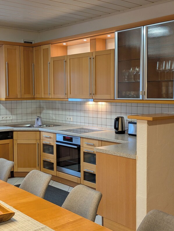 A modern kitchen with wooden cabinets, a built-in oven, a coffee maker, and a dining area with a table and chairs.
