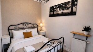 Cama bem arrumada com lençóis brancos e almofada marrom, luminária de cabeceira, radiador e prateleira na parede com uma planta.