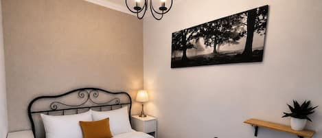 Una cama bien hecha con sábanas blancas y un cojín marrón decorativo, una lámpara de noche, un radiador y una repisa de pared con una planta.