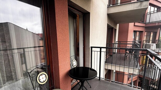 Uma varanda com uma mesa redonda e uma cadeira, uma janela com cortina e vista para edifícios vizinhos.