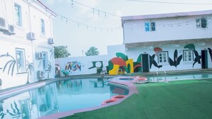 Una piscina con un mural, una zona ajardinada y un edificio con ventanas y unidades de aire acondicionado.