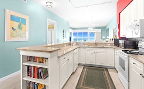 A kitchen with white cabinets, a granite countertop, a built-in bookshelf, and a view of the ocean through large windows. - The Crescent 403 (Miramar Beach)