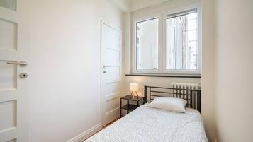 A small, single-bed room with a window, a radiator, and a bedside table with a lamp.