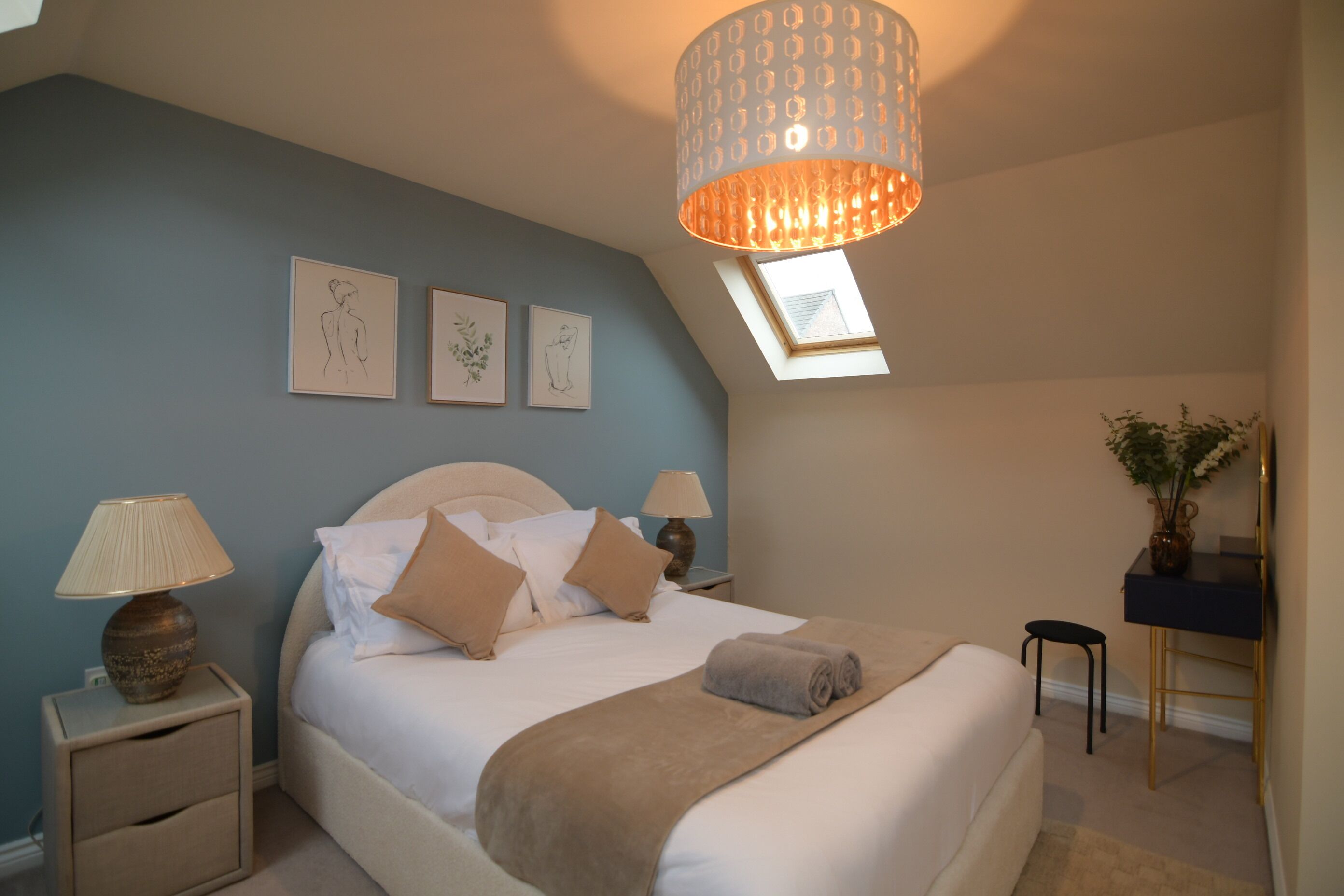 A bedroom with a bed, bedside lamps, a desk, and a skylight.