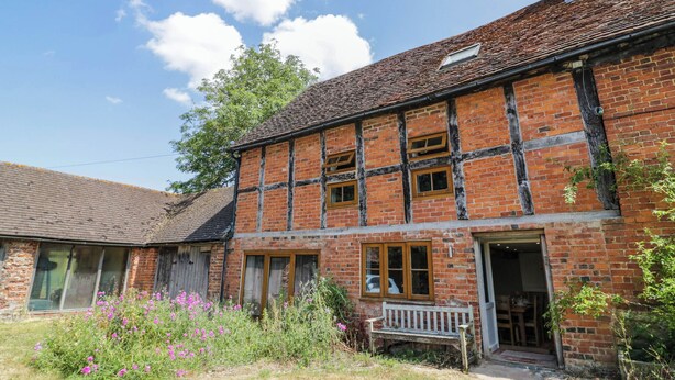A two-story brick building with a sloped roof, a wooden bench outside, and a garden with pink flowers. - Netherend Barn (Malvern)