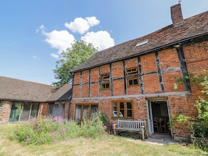 A two-story brick building with a sloped roof, a wooden bench outside, and a garden with pink flowers. - Netherend Barn (Malvern)