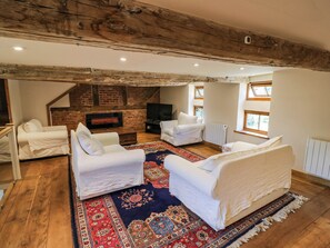 Ein Loft-Wohnzimmer mit freiliegenden Holzbalken, einem Kamin aus Backstein, weißen Sofas und einem großen, gemusterten Teppich.
