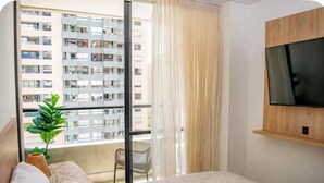 Un dormitorio con cama, televisor montado en la pared, ventana con vista a un edificio y una planta en el alféizar.