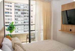 A bedroom with a bed, a TV mounted on the wall, a window with a view of a building, and a plant on the windowsill. - Luxury Apt w/Balcony-Stunning View near el Tesoro (Medellín)