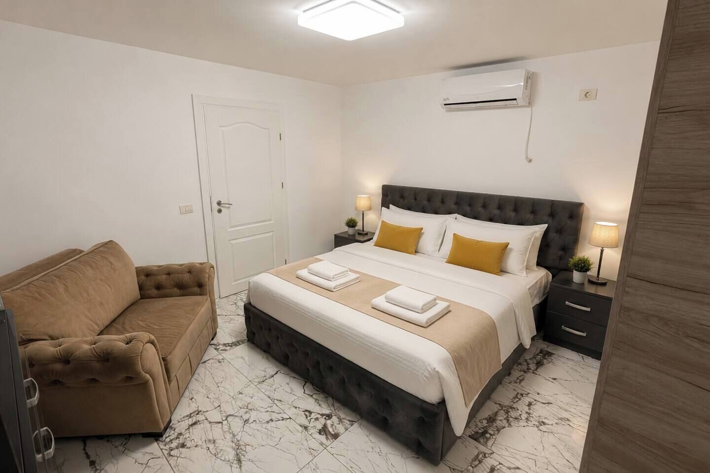 A hotel room with a bed, a sofa, a bedside table with a lamp, and an air conditioning unit on the wall.