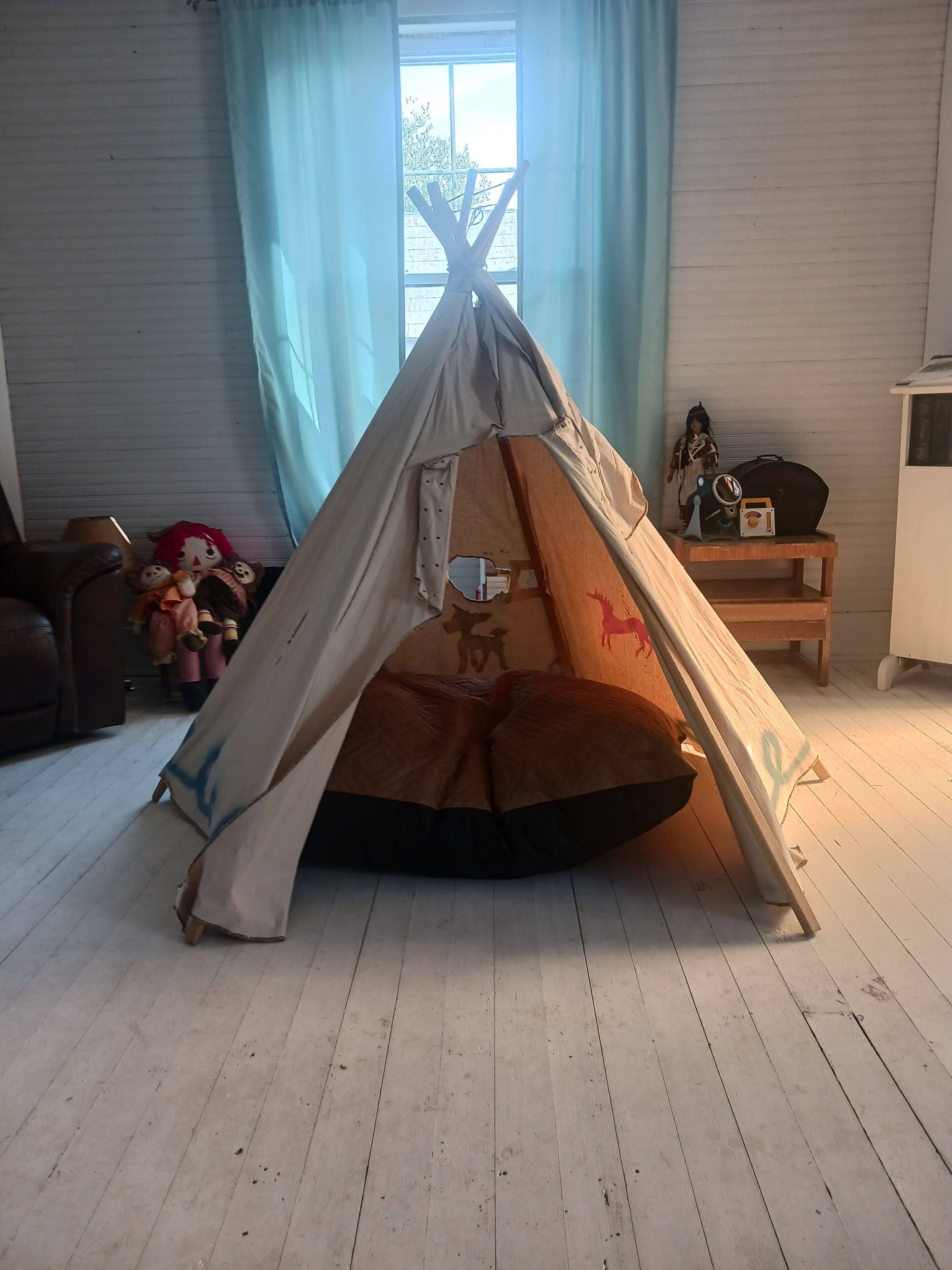 A tent set up in a room with a wooden floor, a window with curtains, and a small table with a doll and a radio.