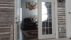 Une porte ouverte donne sur un salon avec une grande fenêtre, un fauteuil en cuir et une décoration murale représentant des bois de cerf.