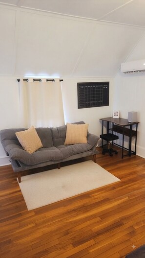 A living room with a grey sofa, two cushions, a small table with a speaker, and a wall-mounted air conditioner. - ​Charming Historic Cozy Getaway • Free Parking! • Walkable to Everything (St. Augustine)