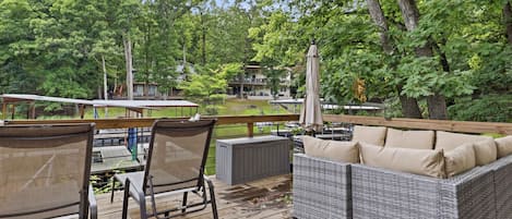 A wooden deck with wicker furniture, a covered table, and a view of a building surrounded by trees.