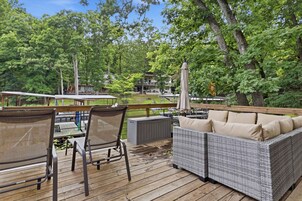 A wooden deck with wicker furniture, a covered table, and a view of a building surrounded by trees.